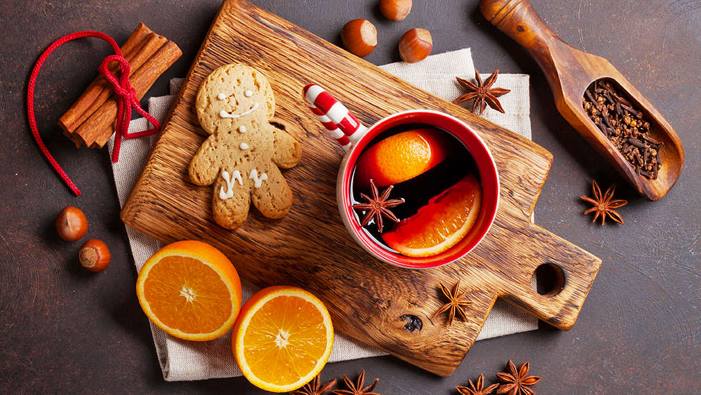 Eine Tasse Glühwein angerichtet auf einem Holzbrett neben einem Lebkuchenmännchen, frischen Orangen und unterschiedlichen weihnachtlichen Gewürzen