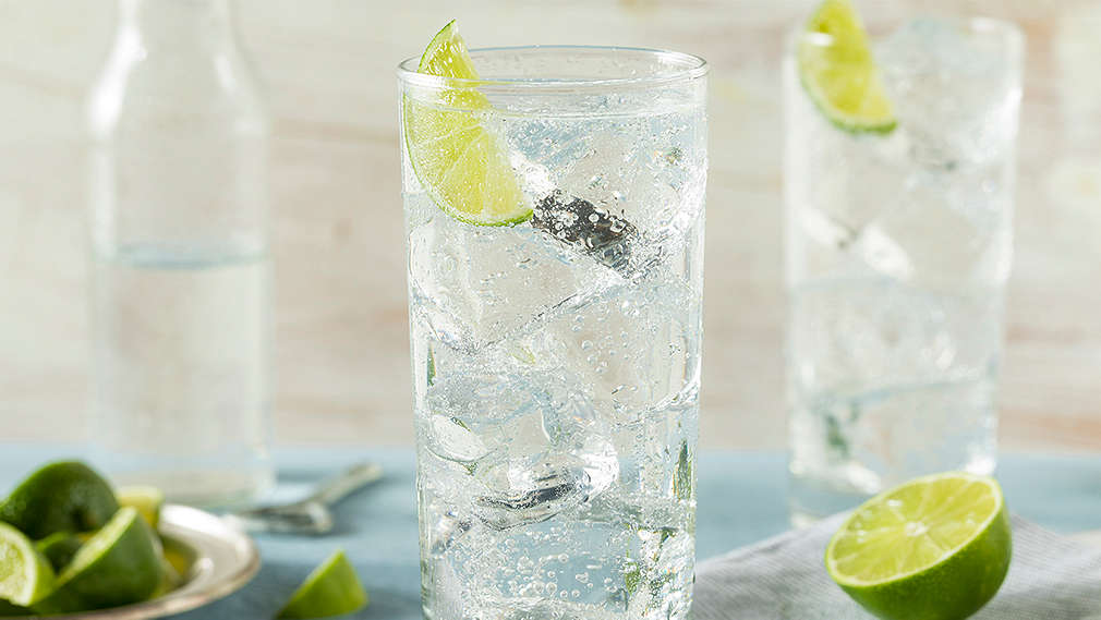 Hellblaues Tischtuch, darauf ein weißes Brett, darauf ein hellblauweißgestreiftes Geschirrtuch, darauf eine halbierte Limette und zwei hohe Gläser mit Eiswürfeln, Sprudelwasser und je einer Scheibe Limette. Im Hintergrund eine halb gefüllte Wasserflasche und ein kleiner Teller mit Limettenstücken. 