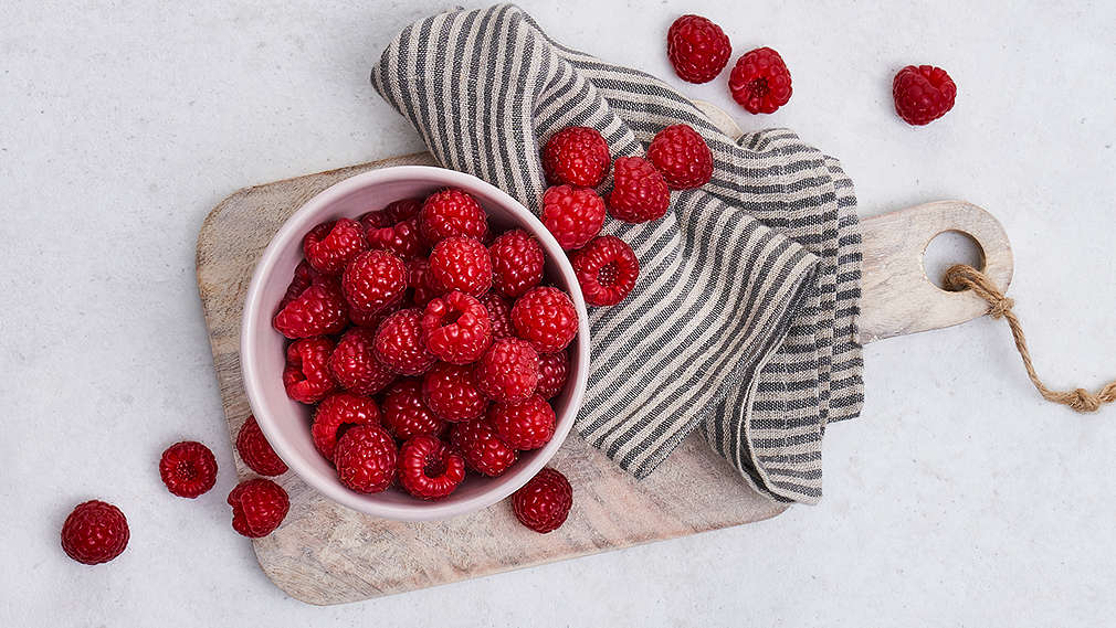 Eine Schale mit Himbeeren steht auf einem Schneidebret auf einem Tisch. daneben liegt ein Küchentuch. 
