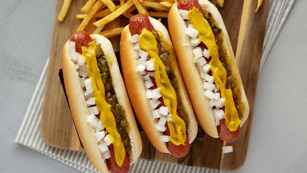 Eine Hand hält einen frischen Hot Dog mit Würstchen und Zwiebelsoße, während die andere Hand den Hot Dog mit Senf aus der Tube toppt