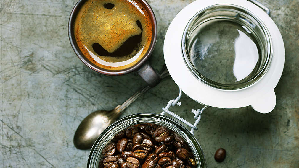 Mehrere Kaffebohnen liegen auf dem Tisch. Ihnen gegenüber steht ein Tasse Kaffee und ein geöffnetes Glas mit Kaffeebohnen 