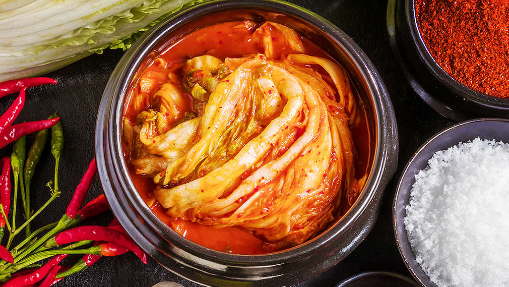 Eine von oben fotografierte dunkle Arbeitsfläche, auf der Chinakohl, kleine Chili-Sträußchen, Knoblauch und kleine Schälchen mit Chilipulver, Chilischoten, Meersalz liegen. In der Mitte ist ein größeres Schälchen, gefüllt mit Kimchi