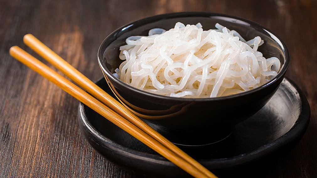 Konjak-Nudeln in einer kleinen schwarzen Schüssel mit schwarzem Untersetzer und Essstäbchen daneben, alles steht auf einem Holztisch