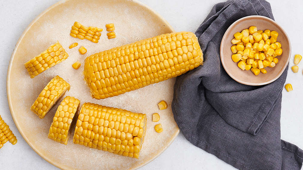 Maiskolben auf einem hellen Teller, daneben eine kleine Schüssel mit einzelnen Maiskörnern auf einem dunkelgrauen Geschirrtuch