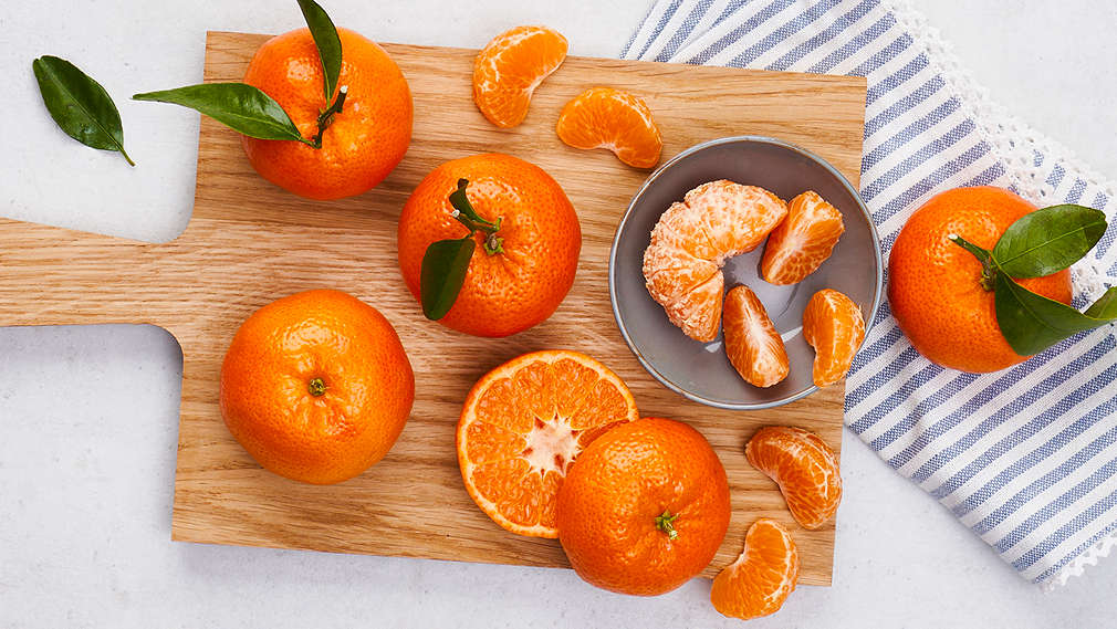 Mehrere Mandarinen liegen auf einem Holz-Schneidebrett auf einem Tisch. Eine davon ist halbiert und eine andere geschält.