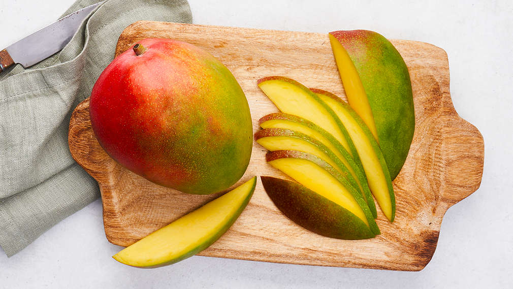 Eine ganze Mango-Frucht liegt auf einem Holzschneidebrett. Links daneben liegt ein Küchenmesser auf einem Küchentuch und rechts auf dem Schneidebrett liegt eine Hälfte einer Mangofrucht in Stücke geschnitten.Tee steht auf einem Tisch. daneben steht eine kleine Schale mit Matcha Tee Pulver, eine Schale mit  dem Matcha tee eis und einem Match Besen.