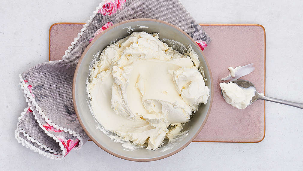 Eine mittelgroße Schale mit Mascarpone steht auf einem Tisch. Daneben liegt ein Esslöffel.