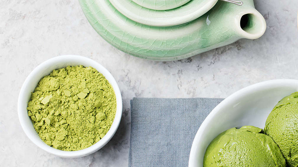 Eine kleine Kanne Matcha-Tee steht auf einem Tisch. Daneben steht eine kleine Schale mit Matcha-Pulver, eine Schale mit Matcha-Eis und einem Match-Besen.