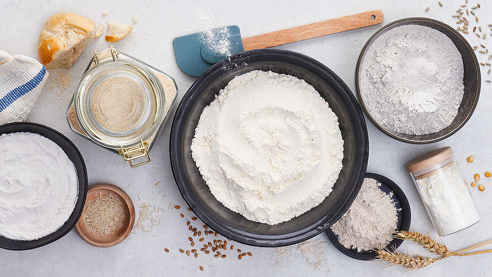 Verschiedene Mehlsorten in Schüsseln neben weiteren Backzutaten, Backzubehör und Getreidesamen