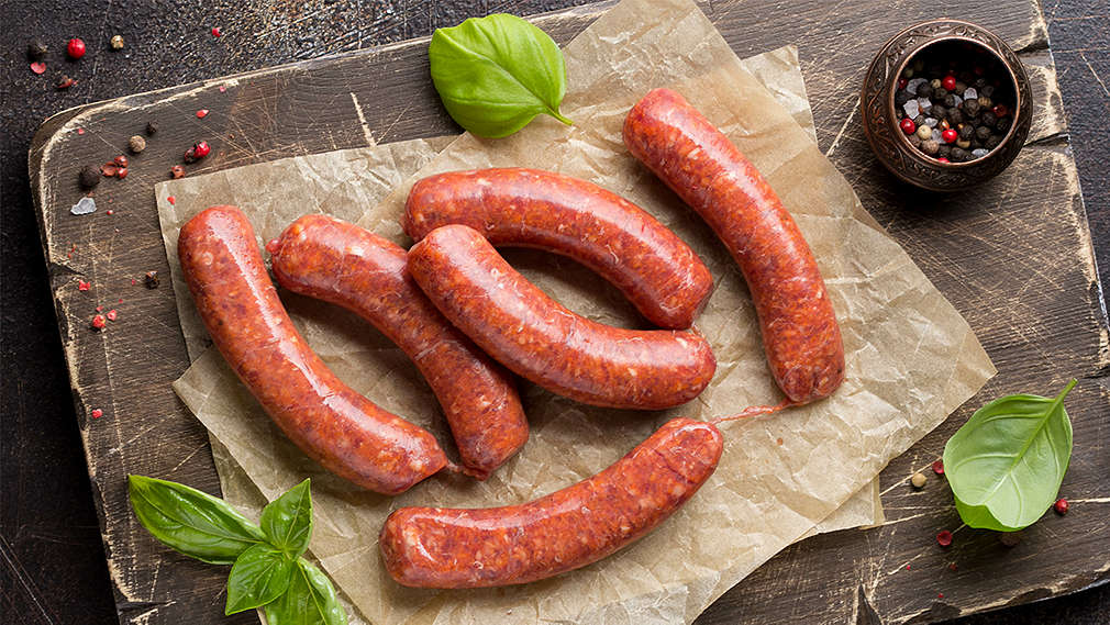 Zu sehen sind mit Basilikum und buntem Pfeffer garnierte Merguez-Würste auf einem Holzbrett