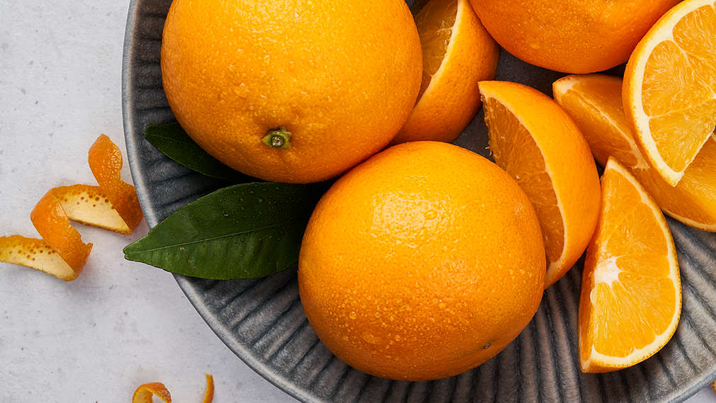 Zwei ganze Orangen, Orangenschnitze sowie Orangenzeste liegen auf einem Teller