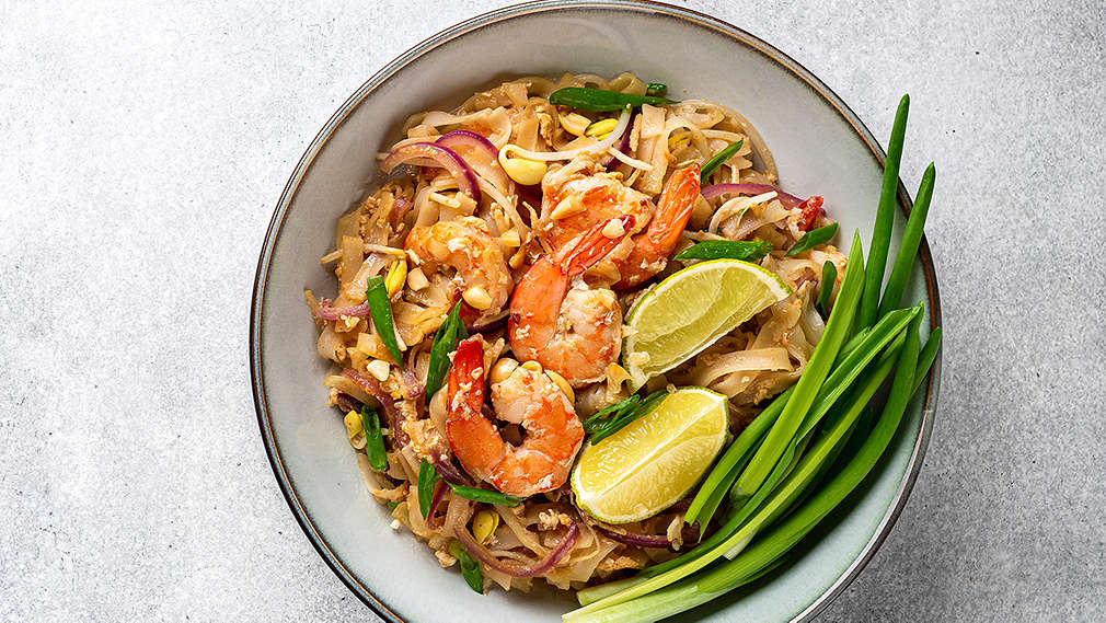 Ein Teller mit frisch gebratenem Pad Thai, das mit Erdnusskernen, Riesengarnelen, Frühlingszwiebeln und einer ausgepressten Limette getoppt ist, vor blauem Hintergrund
