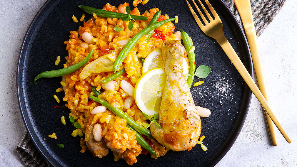 Auf einem hellen Untergrund liegt ein gestreiftes Geschirrtuch. Darauf steht ein schwarzer Teller mit einer Portion Paella. Daneben liegt goldenes Besteck