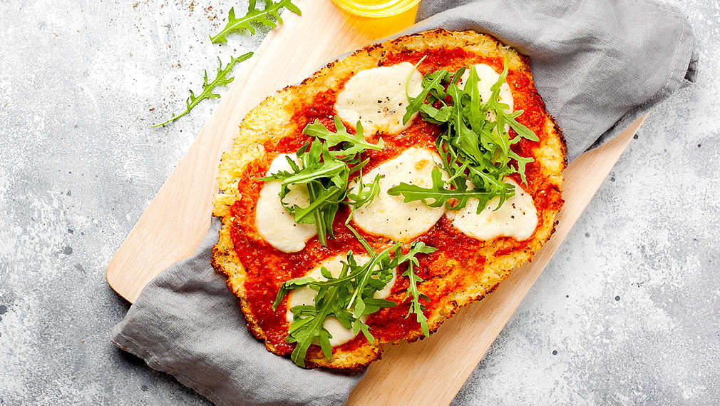 Zu sehen ist eine Pinsa belegt mit Salami, Kresse und Sprossen auf einem runden Holzbrett auf grauer Arbeitsfläche, daneben einige Kirschtomaten