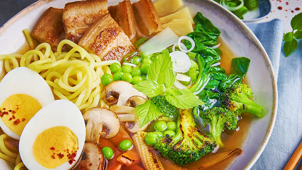 Eine Schüssel gefüllt mit Ramen-Suppe, die mit Toppings wie Schweinebauch, halbierten Eiern, Pilzen und Gemüse angerichtet ist. Daneben rechts liegt ein blaues Serviettentuch und darauf ein Paar Stäbchen