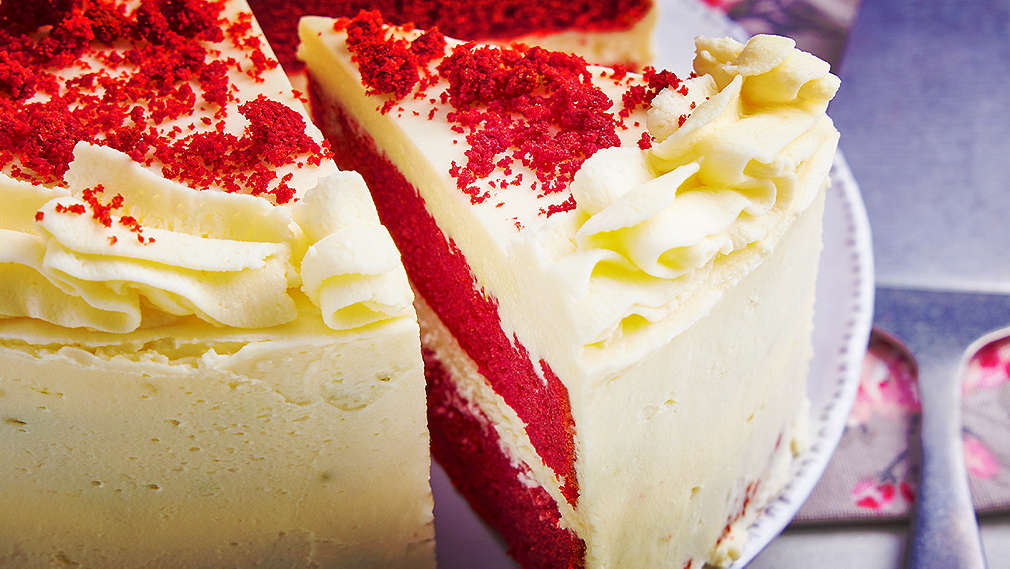 Abbildung eines Red-Velvet-Kuchens im Closeup auf einem Teller, von dem ein großes Stück ausgeschnitten, das leicht herausgezogen ist, sodass man die inneren zwei roten Schichten und die weiße Schicht in der Mitte sehen kann. Rechts neben dem Kuchenteller liegt eine gemusterte Serviette und darauf ein Tortenheber.   
