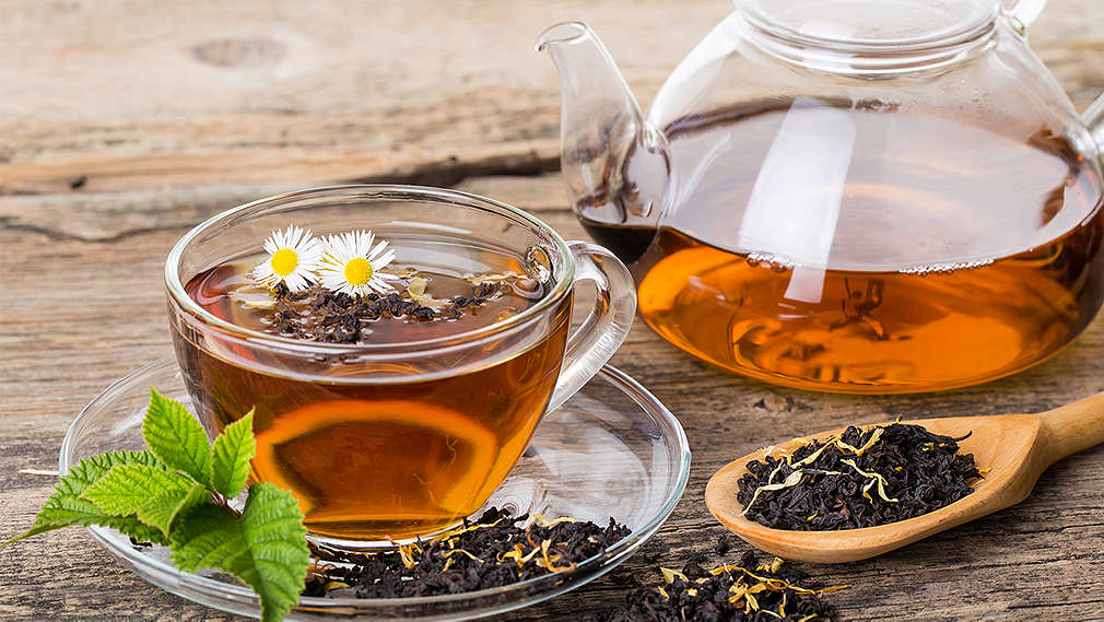 Schwarzer Tee in einer Tasse und einer Teekanne auf Holzuntergrund, rechts daneben liegt ein Löffel mit losem schwarzen Tee 