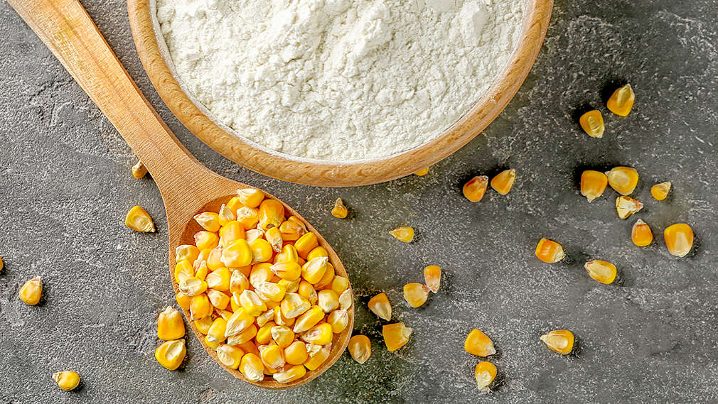 Eine Holzschale gefüllt mit Speisestärke und ein Holzlöffel mit Maiskörnern auf einem grauen Hintergrund. Die Maiskörner sind dazu noch auf dem Bild verstreut.