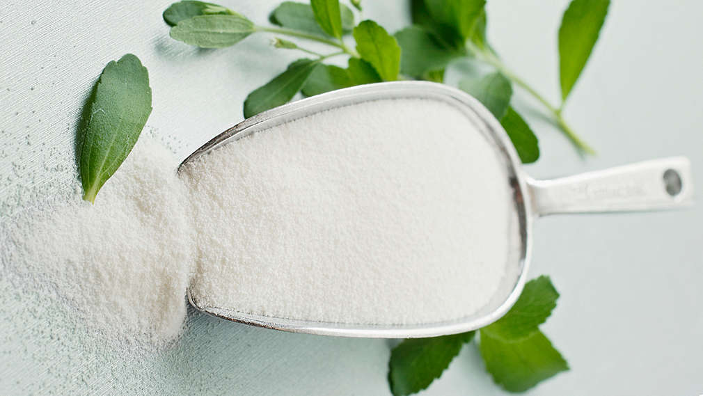 Stevia in Pulverform in Dosierschaufel und daneben frische Steviablätter