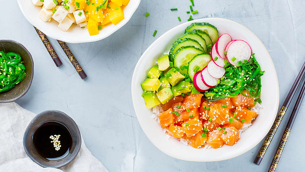 Zu sehen ist eine Sushi Bowl mit Reis, rohen Lachswürfeln, Avocadowürfeln, Gurkenscheiben, Radieschenscheiben und Seetang. Eine weitere Schale ist links im Bild, darin sind Reis, Tofuwürfel, Mangowürfel, rote Zwiebelscheiben, Avocadowürfel und Radieschenscheiben. Außerdem im Bild: zwei Paar Stäbchen und drei Schälchen mit Schnittlauch, Seetang und Sojasauce