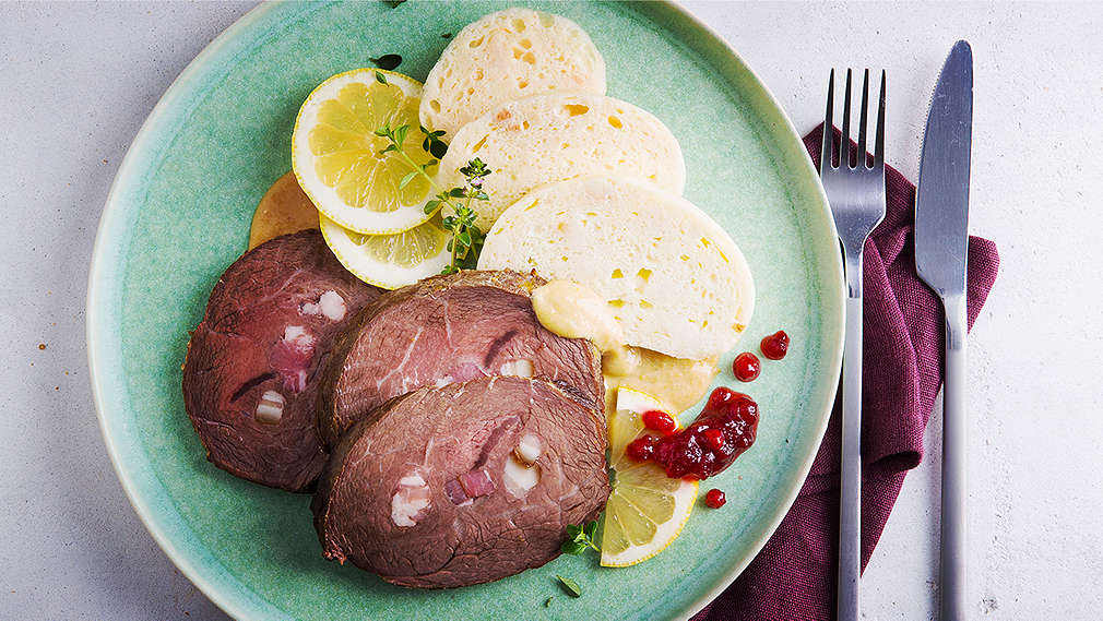Auf hellem Untergrund liegt eine violette Serviette mit silbernem Besteck, daneben ein grüner Teller mit einer Portion Svíčková
