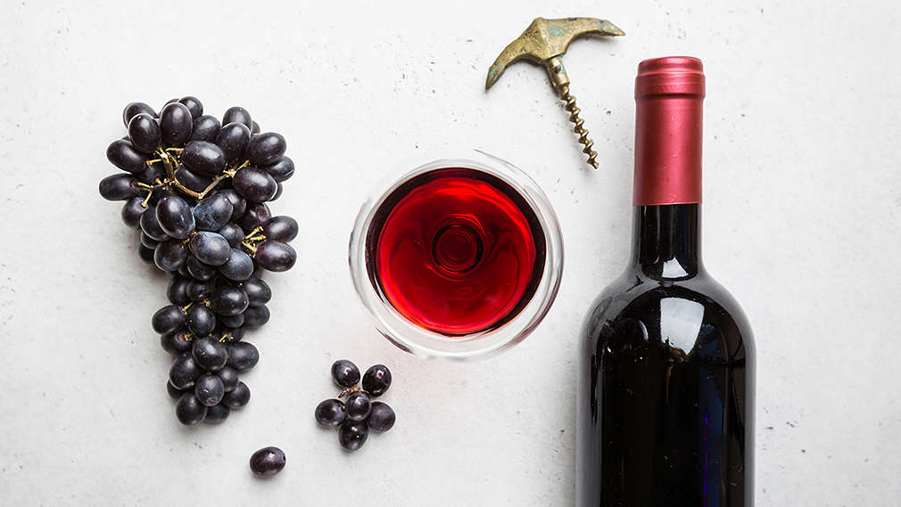 Eine Rispe roter Weintrauben, daneben ein Glas Rotwein sowie eine Rotweinflasche und ein Flaschenöffner von oben fotografiert auf hellgrauem Grund