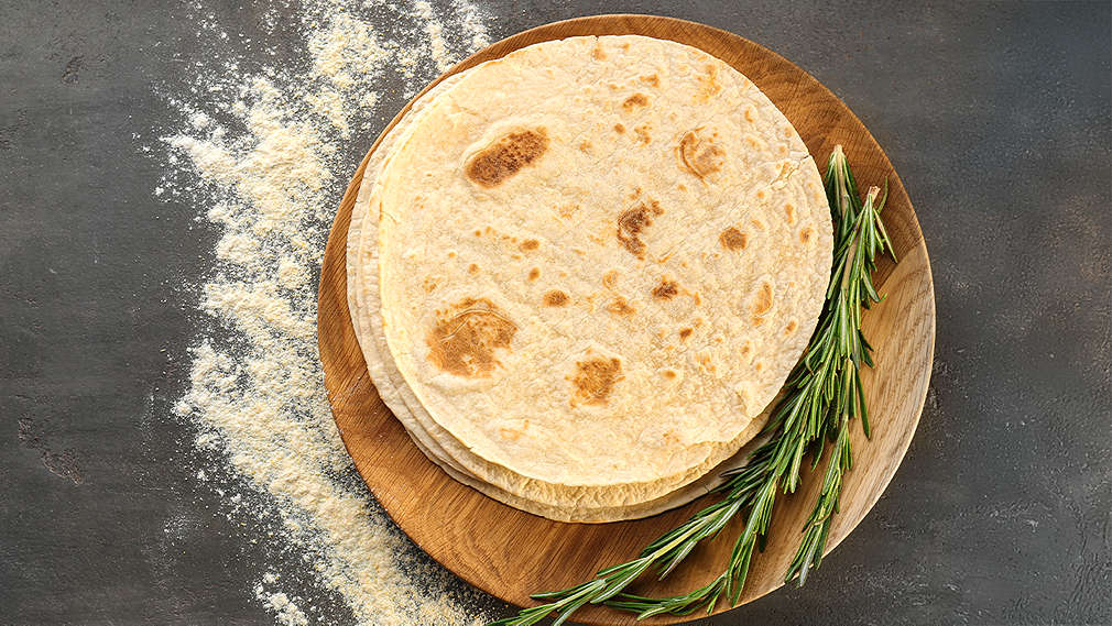 Zu sehen ist ein Stapel mit einigen gelben und schwarzen Tortillafladen in der Hand eines Mannes, der Mann trägt ein graues T-Shirt und eine blaue Schürze