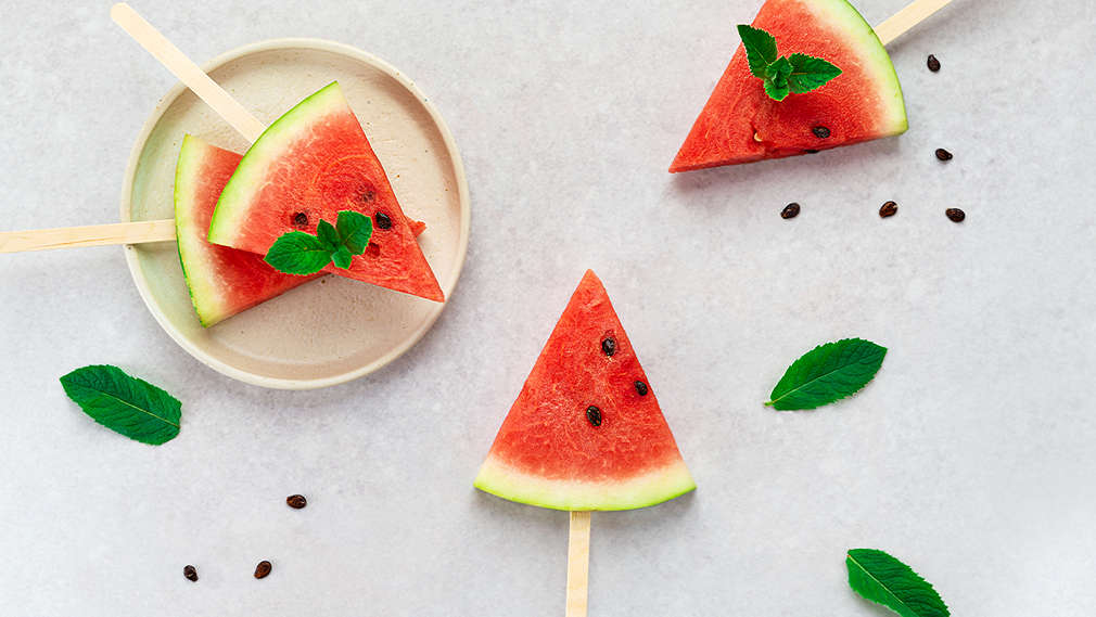 Hellgrauer Untergrund, darauf ein dunkelgraues Geschirrtuch und ein ein braunes Schneidebrett; auf dem Brett liegt eine ganze Wassermelone, aus der eine Scheibe herausgeschnitten wurde, die daneben liegt; außerdem liegen noch ein paar kleinere Stücke der Frucht auf dem Brett