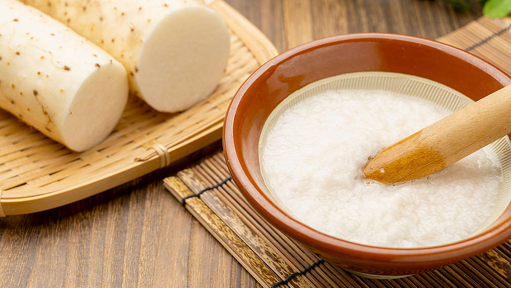 Yamswurzel in ihrer Ursprungsform sowie gemörsert in einer Schale angerichtet auf geflochtenen Tabletts.