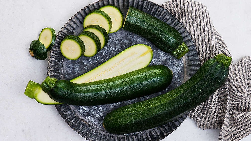 Zwei grüne angeschnittene und eine ganze Zucchini liegen auf einem Teller. 