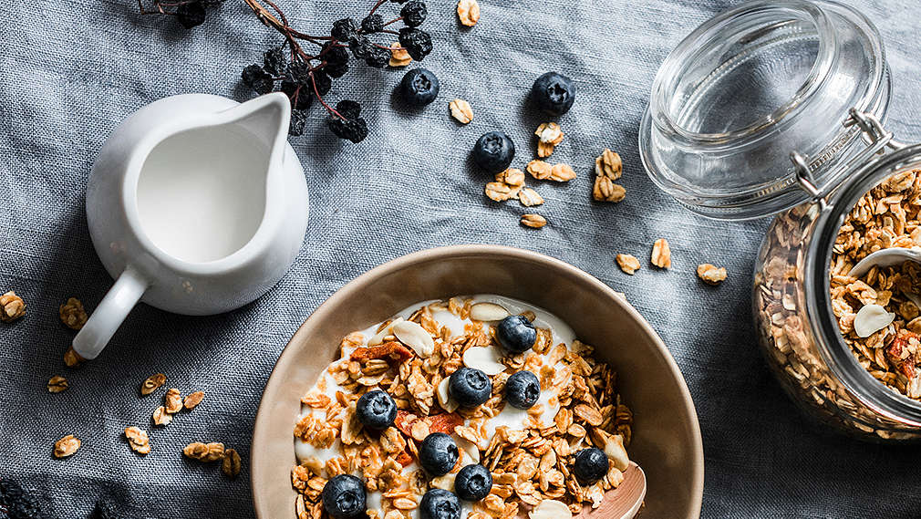 Eine Schale Müsli mit Blaubeeren; daneben ein Kännchen mit laktosefreier Milch