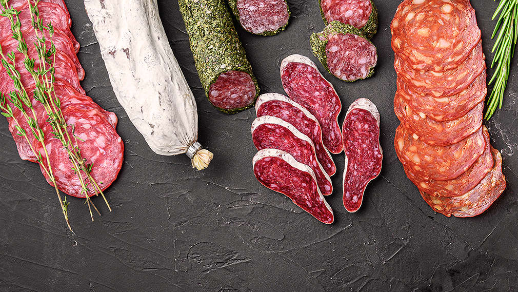 Verschiedene Wurst-Spezialitäten im Ganzen sowie aufgeschnitten, dekoriert mit frischen Gewüzen, auf dunklem Hintergrund.