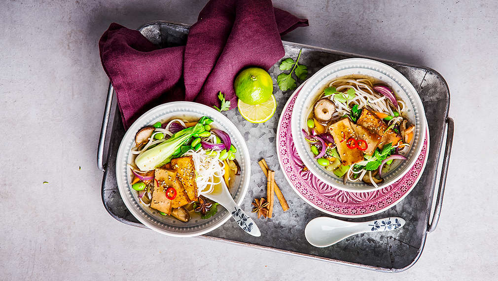 Na szarej emaliowanej tacy stoi azjatycka miseczka z zupą pho z kapustą pak choi, makaronem i plastrami tofu. Na tacy znajdują się druga, pokazana w przekroju miseczka, ściereczka kuchenna w kolorze czerwonego wina, przecięta limonka, dwie laski cynamonu i anyż gwiaździsty.