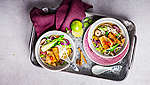 На изображении азиатская миска для супа на серой эмалированной посуде, наполненная супом Фо с бок-чой, лапшой и ломтиками тофу. Кроме того, на подносе видна еще одна миска, красное кухонное полотенце, разрезанный лайм, две палочки корицы и звездчатый анис.