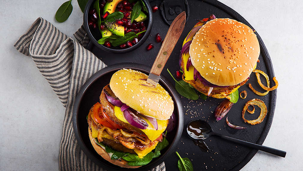 Dva veganské burgery na černém kulatém talíři. Burgery jsou proložené grilovanou zeleninou, mimo jiné cibulemi, rajčaty a špenátem, veganským karbanátkem a veganským sýrem. Vedle toho je v malé misce salát z avokáda a granátového jablka.
