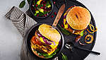 Dva veganska hamburgera na crnom okruglom tanjuru. Hamburgeri su punjeni grilanim povrćem, između ostalog lukom, rajčicom i špinatom, kao i veganskom pljeskavicom i veganskim sirom. Pored se nalazi salata od avokada i sjemenki nara u maloj zdjelici.