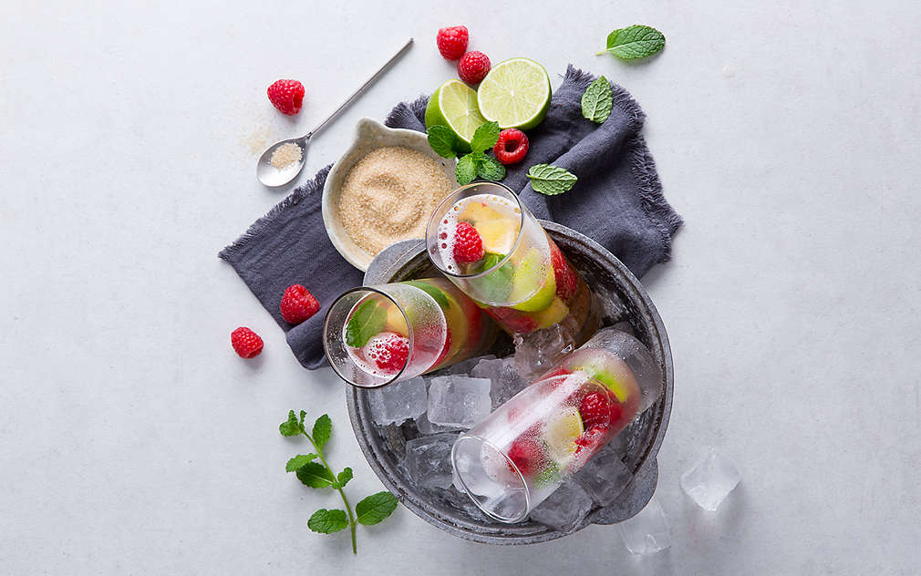 W wiaderku pełnym kostek lodu widać trzy szklanki wypełnione malinami, limonkami, miętą i pomarańczą. W tle widać granatową ściereczkę kuchenną, a na niej miseczkę z cukrem trzcinowym, przepołowioną limonkę, miętę i maliny.