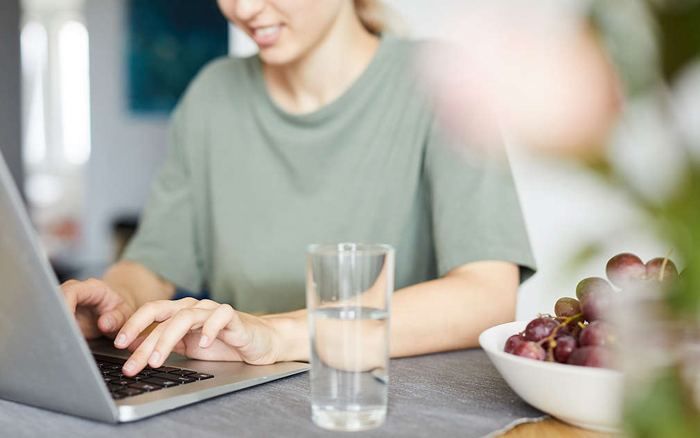 O femeie tânără cu un tricou verde stă la un birou gri și lucrează, având un pahar mare de apă și un bol de struguri lângă ea.