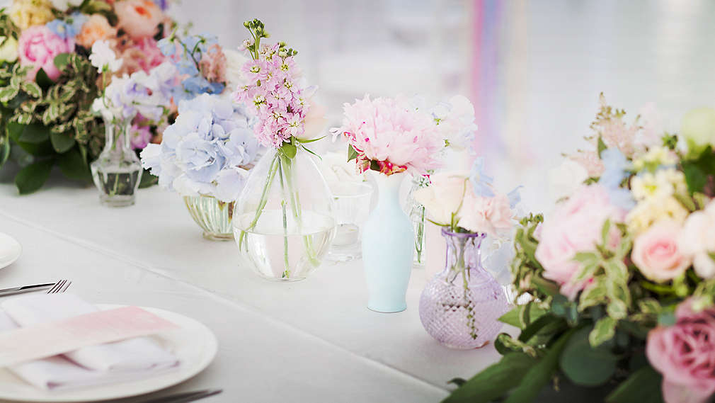 Masă de nuntă cu multe vaze mici, de diferite forme și culori, cu flori în diverse nuanțe pastelate.
