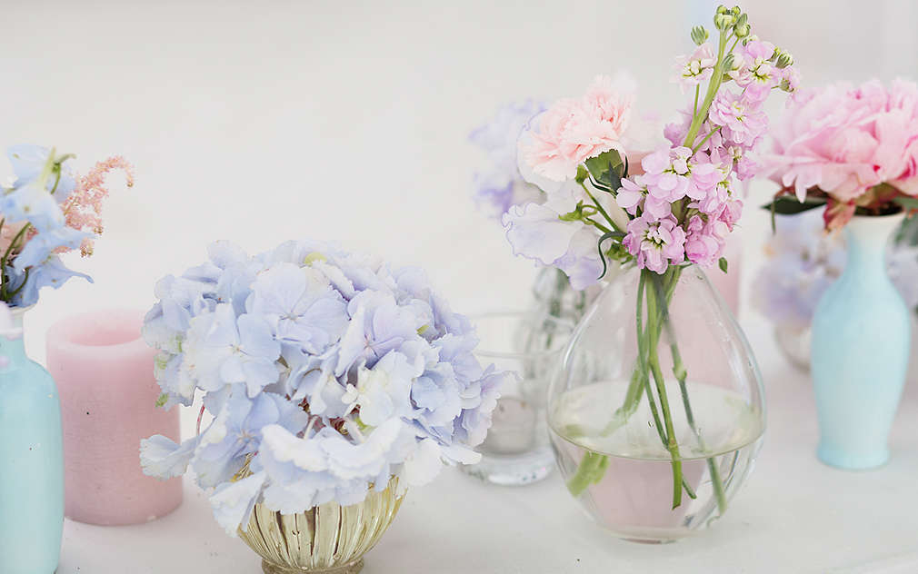 Stół weselny w kolorze białym, na którym ustawione są różne małe wazoniki z kwiatami, wykonane ze szkła i ceramiki.