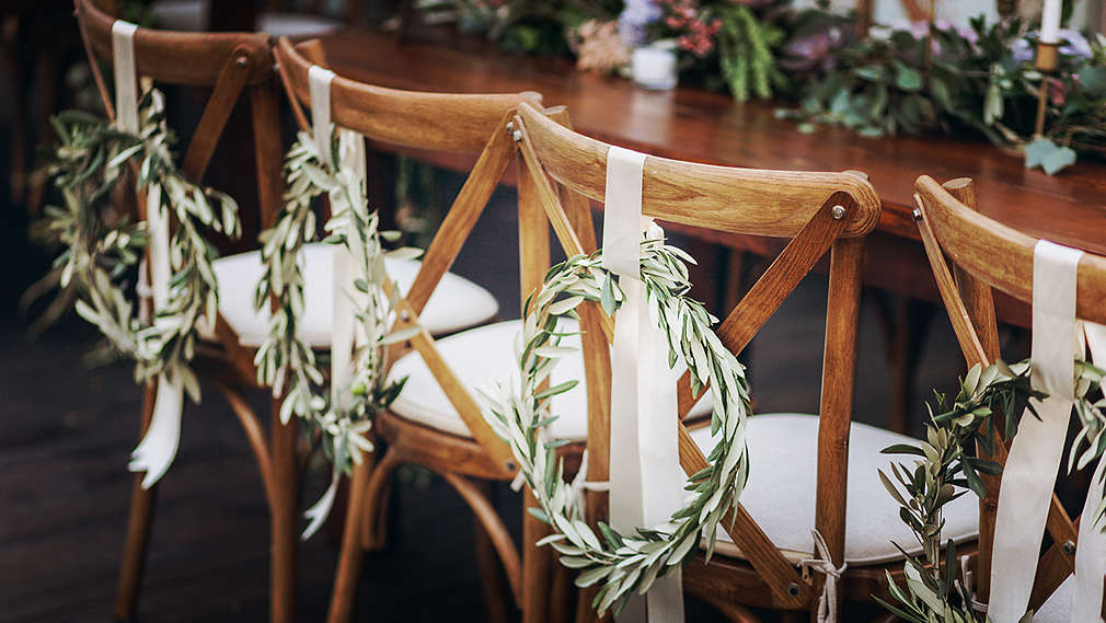 Stół weselny, przy którym stoją drewniane krzesła z białymi poduszkami na siedzeniach, każde ozdobione wiankiem z gałązek przymocowanych do białej kokardy.