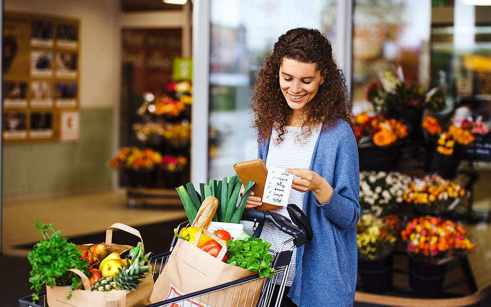 Na obrázku je žena s plným nákupným vozíkom, ktorá práve vychádza z predajne Kaufland a kontroluje si nákupný zoznam.