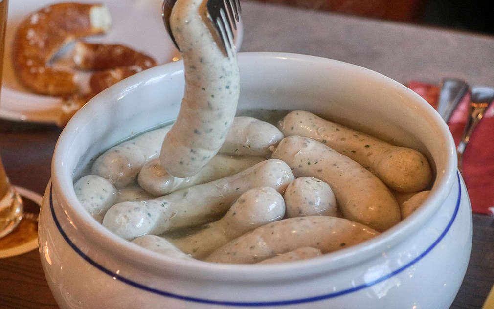 Biele klobásy plávajú vo vode v bielej keramickej mise s typickým bavorským bielo-modrým vzorom. Na pozadí možno rozpoznať košík s praclíkmi a biely tanier, ktorý má takisto modro-biely vzor.