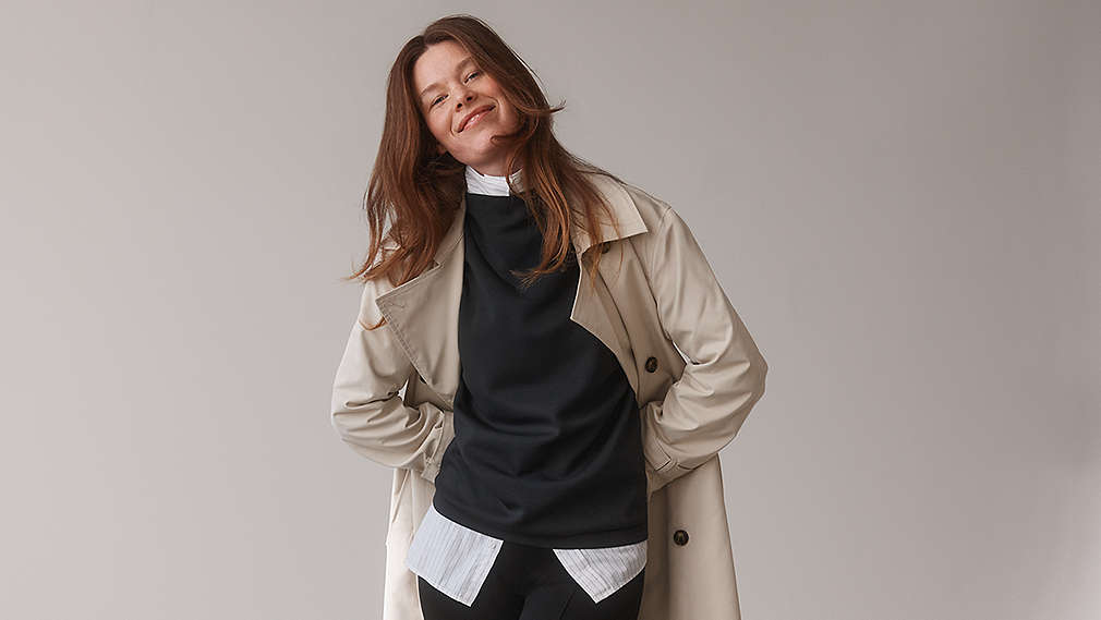 Eine lächelnde Frau mit langen, braunen Haaren präsentiert einen mehrlagigen Look aus einem hellen Trenchcoat, einem schwarzen Oberteil und einer darunter hervorstehenden, gestreiften Bluse., Eine l�chelnde Frau mit langen, braunen Haaren pr�sentiert einen mehrlagigen Look aus einem hellen Trenchcoat, einem schwarzen Oberteil und einer darunter hervorstehenden, gestreiften Bluse.
