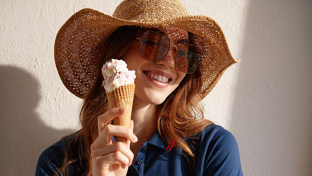 Eine lächelnde junge Frau mit einem Strohhut und einer Sonnenbrille hält eine Waffel mit zwei Kugeln Eis in der Hand., Eine l�chelnde junge Frau mit einem Strohhut und einer Sonnenbrille h�lt eine Waffel mit zwei Kugeln Eis in der Hand.