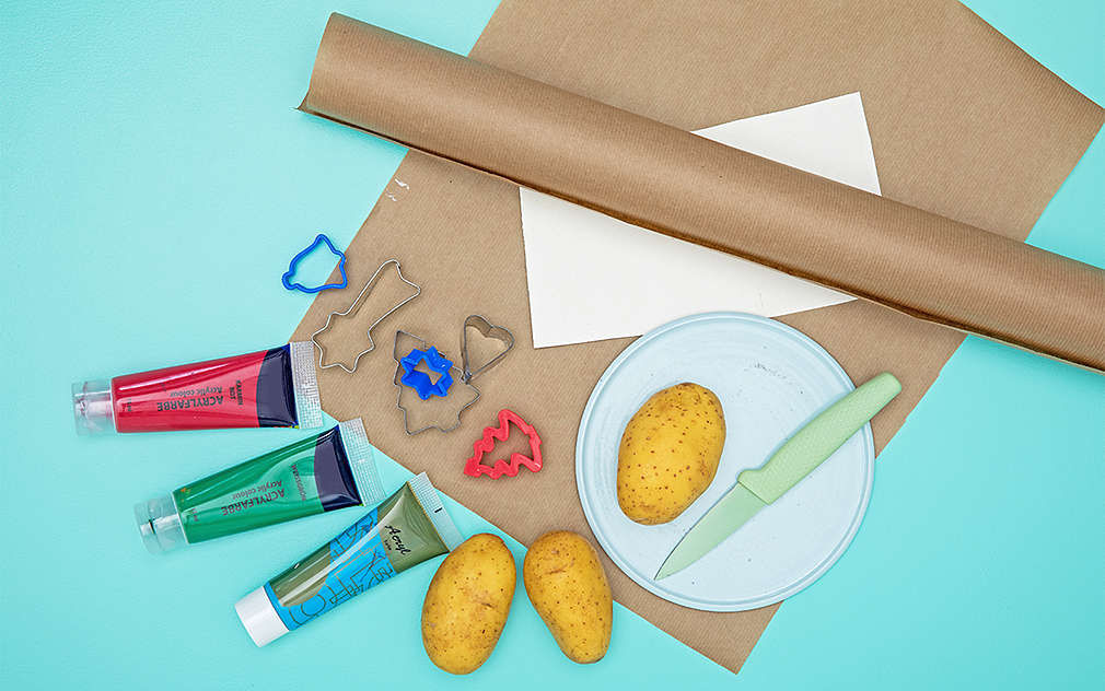 Na turkusowym tle widać z góry papier do pakowania. Na nim są położone talerz z surowym ziemniakiem i nożem, a obok kolorowe farbki, dwa ziemniaki i foremki do ciastek.