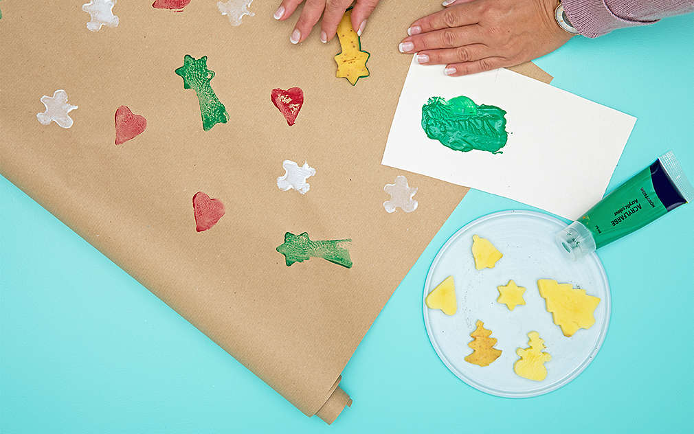 Dwie dłonie używają ziemniaków z wyciętymi motywami jako kolorowych stempli do zdobienia papieru do pakowania.