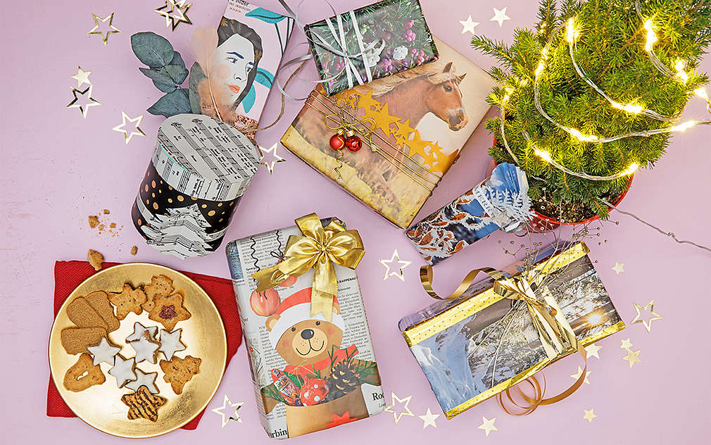 Na różowym tle znajdują się prezenty w różnych opakowaniach. Po lewej i prawej stronie zdjęcia widoczne są miseczka z ciastkami i mała choinka.