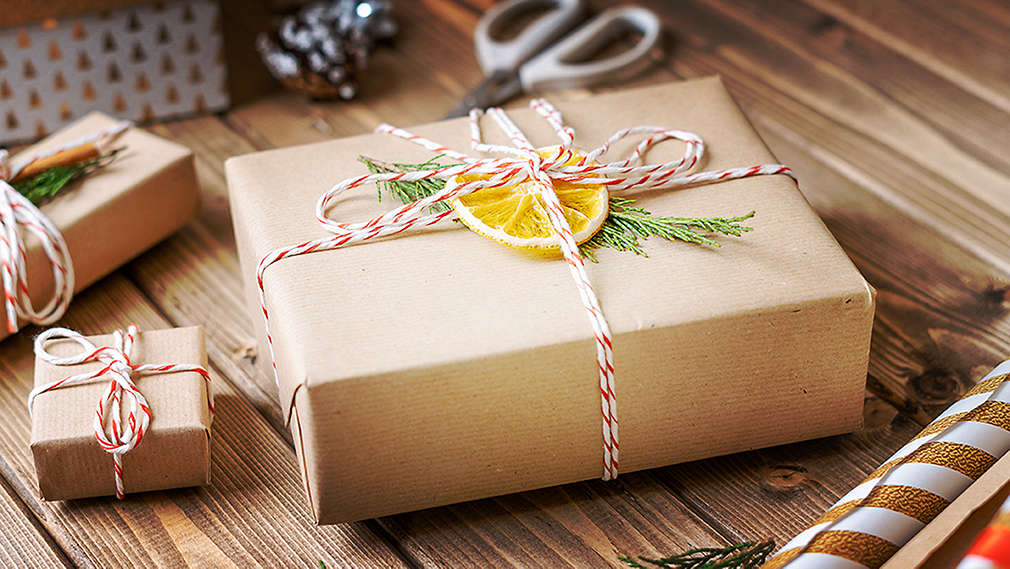 Pe un suport de lemn se află un cadou învelit în hârtie de împachetat, legat cu o panglică roșie și albă și decorat cu o felie de portocală și o ramură de brad. În jurul cadoului se află un cadou mai mic și ustensile pentru împachetat.